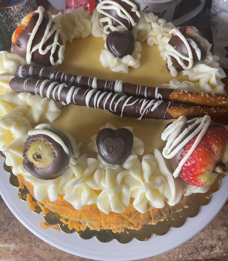 A dense, creamy New York Style Cheesecake Topped with custom chocolate pretzels, chocolate-covered strawberries, cream cheese icing, and a pretzel crust. A heavenly rich classic dessert.